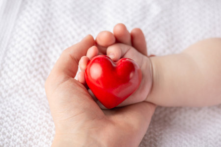 A loving moment as a parent gently holds a newborn baby's hand around a bright red heart, symbolizing care, warmth, family, and maternity in a gentle sceneの写真素材