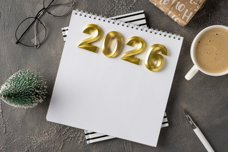 A stylish desk scene features a white notebook, gold 2026 numbers, a warm cup of coffee, a tiny...