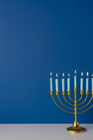 Gold vertical menorah with lit candles sits on a white table against a deep blue backdrop, capturing Hanukkah warmth, tradition, and festive mood suitable for holiday greeting and celebration imageryの写真素材