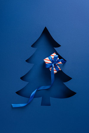 Blue vertical card features a cutout Christmas tree with a striped gift tied by a blue ribbon, ideal for holiday greetings and festive themes.の写真素材