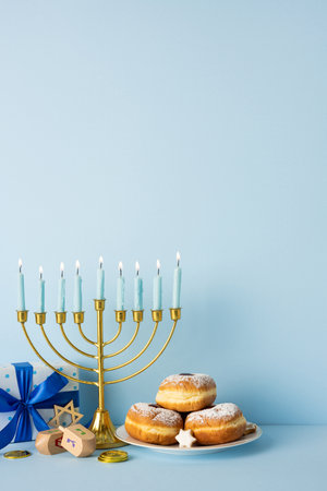 A vertical festive Hanukkah scene featuring a glowing menorah, powdered donuts, a plate of treats, a dreidel, coins, and blue wrapped gifts set against a soft blue backdropの写真素材