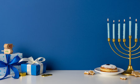 Vibrant Hanukkah display with a golden menorah, blue wrapped gift, dreidel, and festive cookie on a white surface against deep blue wall. A moment of tradition, celebration, gift, and seasonal warmthの写真素材