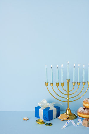 A vertical festive Hanukkah display features a gold menorah, blue gift box, star shaped cookie, donuts, coins and decorative ribbons on a soft blue backgroundの写真素材