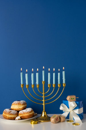 A vertical festive Hanukkah scene with a lit menorah, soft candle glow, donuts and coins, and wrapped gifts on a blue backdrop. Warm, celebratory mood suitable for holiday greetingsの写真素材