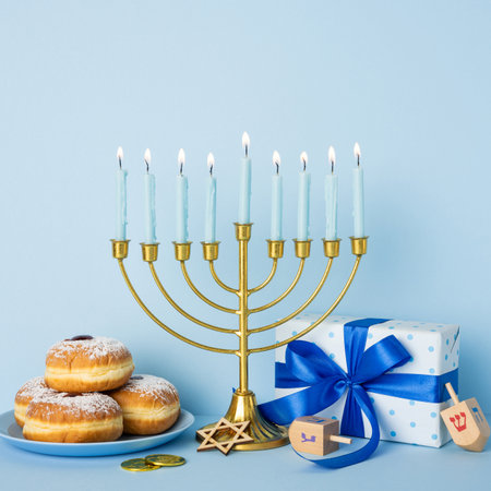 A festive Hanukkah scene features a lit menorah, donuts with powdered sugar, and a blue gift box with a ribbon, conveying tradition, warmth, and holiday greetingsの写真素材
