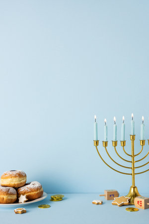A vertical warm Hanukkah scene featuring a gold menorah lit with candles, donuts, coins, and cookies on a pale blue backdrop, conveying festive greeting and holiday traditionの写真素材