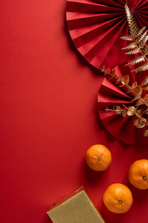 Vibrant vertical red background highlights Chinese New Year mood featuring decorative paper fan gold accents oranges gift box and dried foliage symbolizing luck prosperity and festive greetingの写真素材