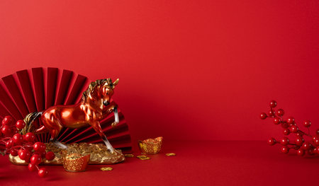 Side view of a vivid red backdrop featuring a golden horse statue, gold ingots, coin treasure, and a red fan, capturing festive luck and prosperityの写真素材