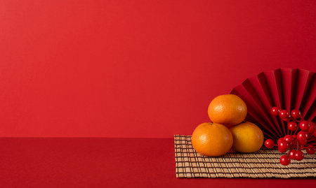 Side view of a festive red backdrop with oranges stacked on a woven mat, red fan, and berries, symbolizing luck and celebration for Chinese New Year greetingの写真素材