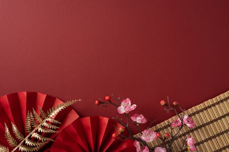Bright red background with decorative paper fans, fern fronds, pink blossoms and a woven bamboo mat create a festive scene for Chinese New Year greetingの写真素材