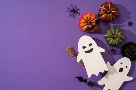 Happy Halloween flat lay featuring paper ghost cutouts, decorative pumpkins, bats and spiders arranged on a purple background to evoke spooky, festive autumn mood