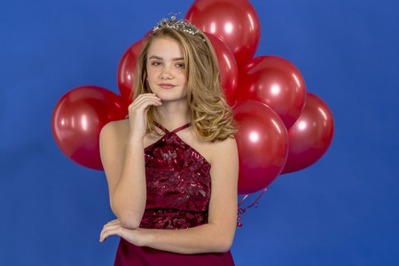 A beautiful blonde teenage model posing in a tiara and red balloons in front of the camera in a studio environmentの写真素材