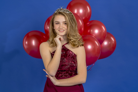 A beautiful blonde teenage model posing in a tiara and red balloons in front of the camera in a studio environmentの写真素材