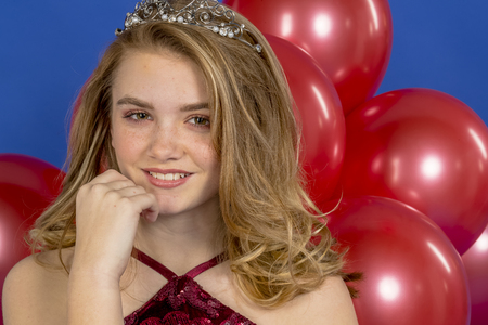 A beautiful blonde teenage model posing in a tiara and red balloons in front of the camera in a studio environmentの写真素材
