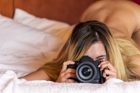 A gorgeous blonde model poses nude while taking photos with a dslr camera in a home environmentの写真素材