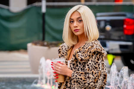 A gorgeous young blonde model poses outdoors while enjoying a winters day in a city environment drinking her coffeeの写真素材