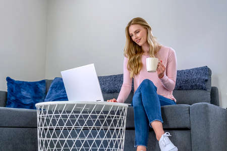 Nice beautiful woman with blonde hair works on the laptop as she sits down on the sofa at homeの写真素材