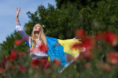 A young blonde Ukrainian woman stands in a field of Red Poppy flowers holding the flag of Ukraine showing her support for the war in her native country of Ukraineの写真素材