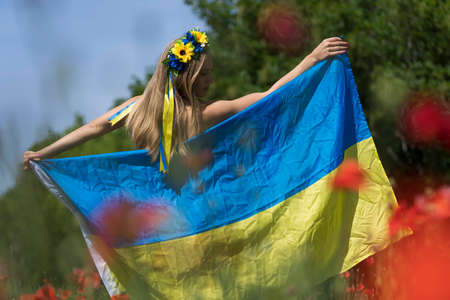 A young blonde Ukrainian woman stands in a field of Red Poppy flowers holding the flag of Ukraine showing her support for the war in her native country of Ukraineの写真素材