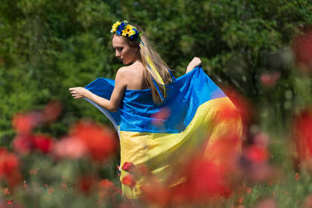 A young blonde Ukrainian woman stands in a field of Red Poppy flowers holding the flag of Ukraine...