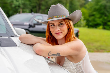 A beautiful redhead model poses in a country setting wearing cowgirl attire against her truck.