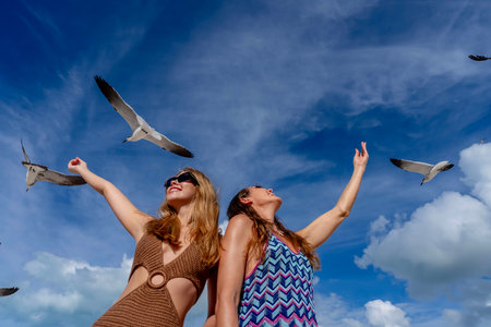 Two stunning women gracefully feed seagulls on a Caribbean beach, surrounded by turquoise waters and golden sands, creating a picturesque vacation sceneA stunning woman gracefully feeds seagulls on a Caribbean beach, surrounded by turquoise waters and golden sands, creating a picturesque vacation sceneの写真素材