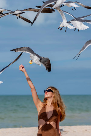 A stunning woman gracefully feeds seagulls on a Caribbean beach, surrounded by turquoise waters and golden sands, creating a picturesque vacation sceneの写真素材