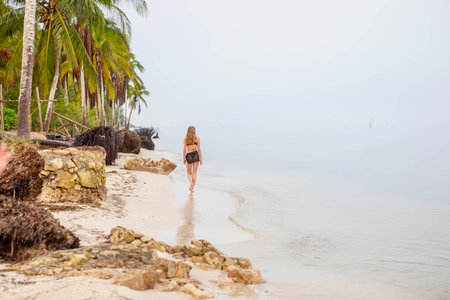 Alone on a Caribbean beach, a stunning woman strolls the fog-kissed coastline, embodying solitude and beauty in the tranquil embrace of a solo vacationの写真素材