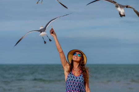A stunning woman gracefully feeds seagulls on a Caribbean beach, surrounded by turquoise waters and golden sands, creating a picturesque vacation sceneの写真素材