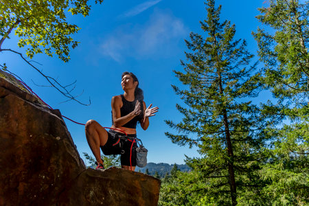 A stunning mixed-race brunette fitness model ascends a rugged mountain trail in the Pacific Northwest, her toned physique glowing in the early afternoon sunlight, surrounded by lush greenery and towering peaks.の写真素材