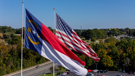 The American flag and North Carolina state flag flutter side by side in the crisp autumn air, vibrant reds, whites, blues, and golds glowing under a clear sky, framed by trees with colorful fall foliage.の写真素材