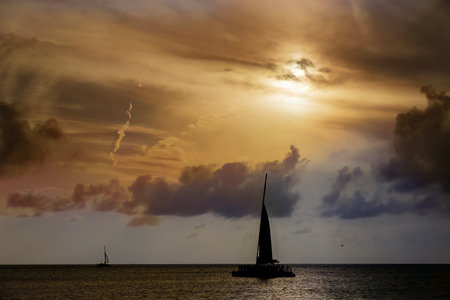 A radiant golden sunset casts shimmering light across calm waters. Silhouetted sailboats gently drift, their dark forms contrasting with the fiery hues of the sky, evoking serenity and wonder.の写真素材