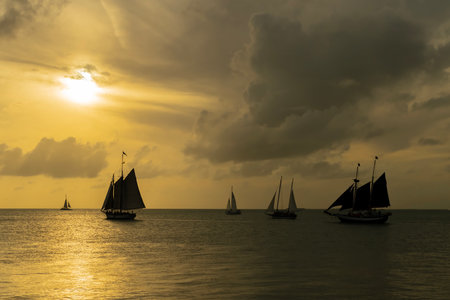 A radiant golden sunset casts shimmering light across calm waters. Silhouetted sailboats gently drift, their dark forms contrasting with the fiery hues of the sky, evoking serenity and wonder.の写真素材
