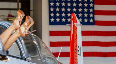 A stunning blonde model radiates retro charm, posing in classic Americana attire alongside a vintage WWII aircraft, framed by a giant American flag. Her throwback U.S. Air Force look exudes patriotic style.の写真素材