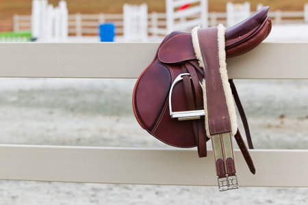 A close-up of a rider's stirrup and English saddle resting on a fence, ready for a lesson. Leather gleams in the light, reins coiled neatly, anticipation hanging in the quiet barn air.