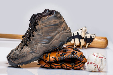 A worn-out pair of dirty baseball cleats sit in a studio setting with a scuffed glove, weathered bat, used batting gloves, and a baseball, all on a clean white backdrop telling tales of the game.の写真素材