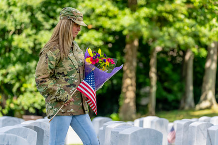 A young woman kneels at a military cemetery amid uniform rows of graves. Clutching flowers and a flag, tears streak her face as she mourns a beloved family member with profound, silent grief.の写真素材