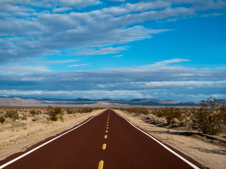 A long, endless road stretches through the vast Mojave Desert, framed by blue skies and scattered white puffy clouds, evoking freedom, solitude, and the beauty of open landscapes.の写真素材