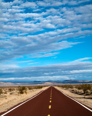 A long, endless road stretches through the vast Mojave Desert, framed by blue skies and scattered white puffy clouds, evoking freedom, solitude, and the beauty of open landscapes.の写真素材