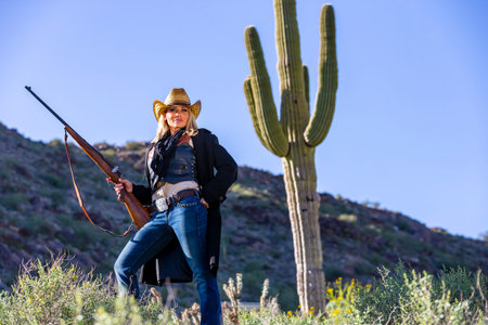 A blonde model in cowgirl gear poses with a rifle in the American Southwest, framed by rugged desert scenery, wide-brim hat, and boots, radiating rustic charm and western spirit.の写真素材