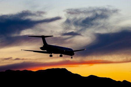 A commercial aircraft glides down at sunset, silhouetted against a sky ablaze with orange, pink, and...