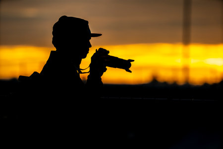 A photographer stands silhouetted against a glowing sunset, camera raised, capturing the vibrant sky as warm hues of orange and pink stretch across the horizon.の写真素材