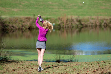 A young woman savors warm spring air as she tees off on lush fairways, smiling in sunlight, confident swing flowing with the breeze, enjoying every graceful moment of her golf round.の写真素材