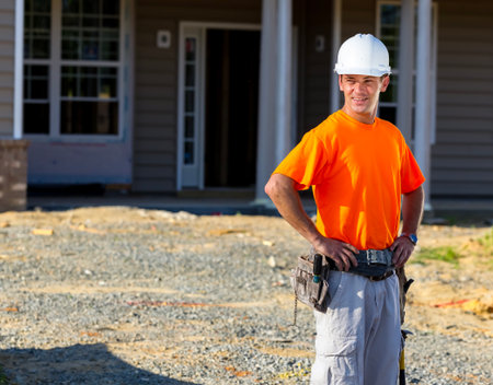 At a sunlit job site, a lead construction worker stands before a rising family home, studying...