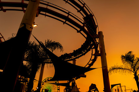Silhouette of a roller coaster rises against a glowing sunset on the California coastline, looping tracks outlined in gold and purple as the ocean reflects the fading light.の写真素材