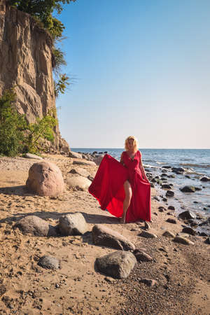 Creative holiday seaside session. Beautiful woman on the seashore in an elegant red dress.の写真素材