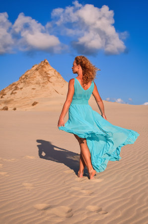 Beautiful blonde woman on hot sand with blue sky in the background. Photo taken on the shifting dunes over the Baltic Sea in Leba, Poland. Photo with a shallow depth of field with blurred background.の写真素材