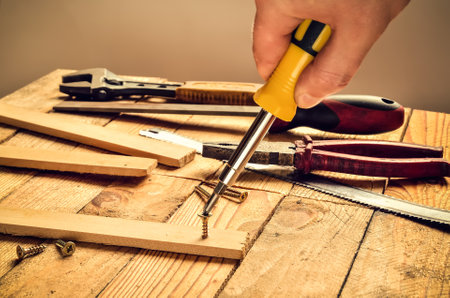Hand holding tool in the workshop. Screwdriver, flat file, pliers, , monkey wrench, screws and boards on natural wooden background.の写真素材