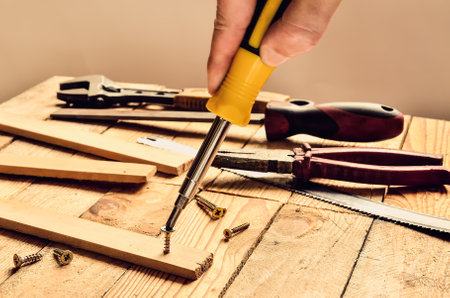 Hand holding tool in the workshop. Screwdriver, flat file, pliers, monkey wrench, screws and boards on natural wooden background.の写真素材