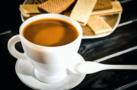 Coffee in a cup with cookies on a plate in the background. Coffee with waffles on a black wooden table. Photo with a shallow depth of field.の写真素材